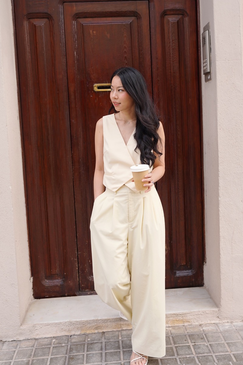 Christine Tan leaning against a wooden door holding a coffee, in a cream waistcoat and trousers